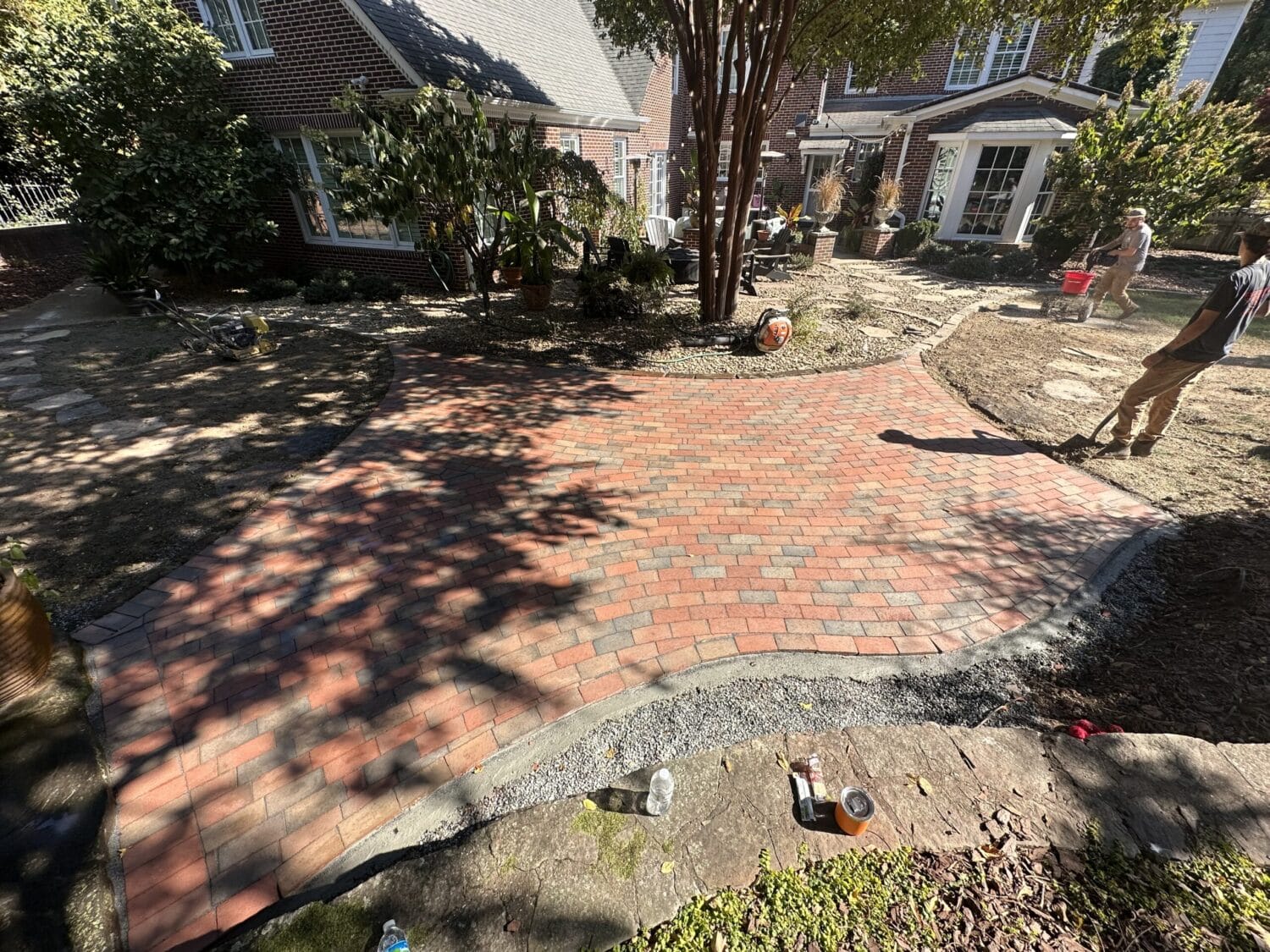 A newly installed red brick patio with curved edges in a residential backyard, surrounded by dirt and construction materials. Two people are working nearby.