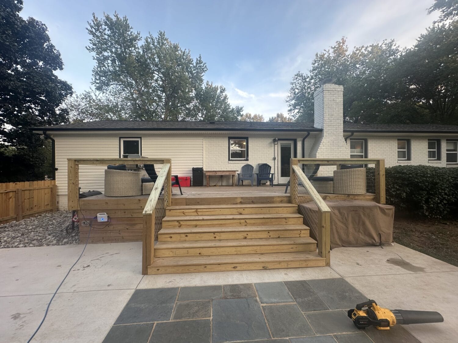 A wooden deck with stairs leads to a white brick house; two round planters, chairs, a table, and covered objects are on the deck. A leaf blower rests on stone pavers in the foreground.