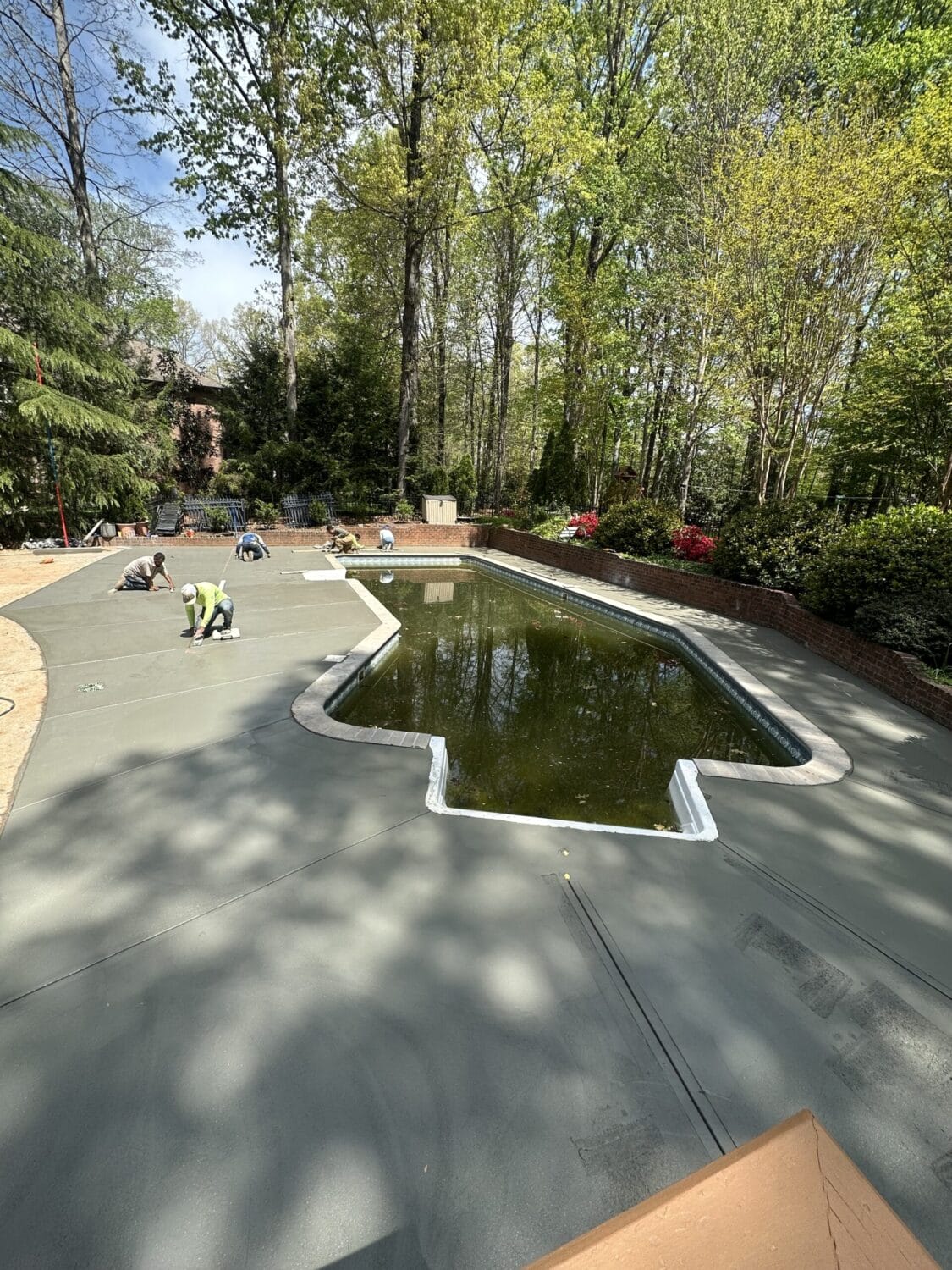 Workers are resurfacing the concrete deck around a partially filled swimming pool in a landscaped backyard surrounded by trees and shrubs.