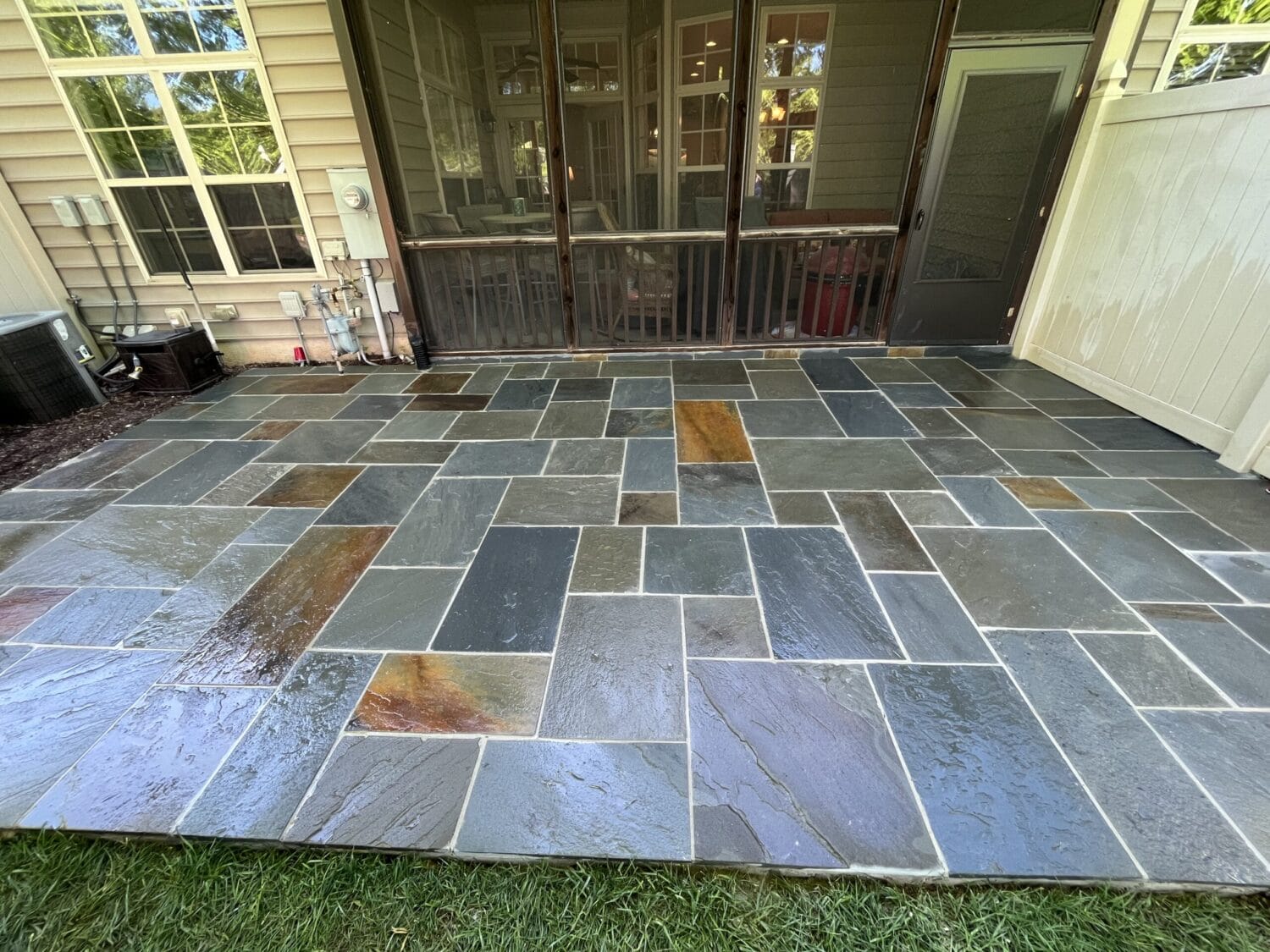 A newly installed outdoor patio features wet, multicolored rectangular stone tiles in various sizes, positioned next to a screened porch and white privacy fence.