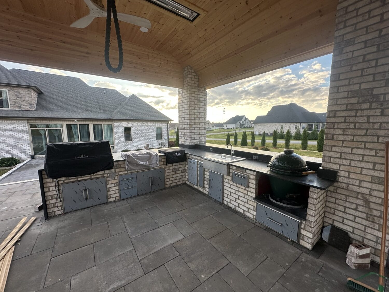 Outdoor kitchen with brick and stainless steel appliances, including a grill and a green ceramic smoker, under a wooden ceiling with a view of neighboring houses and a cloudy sky.