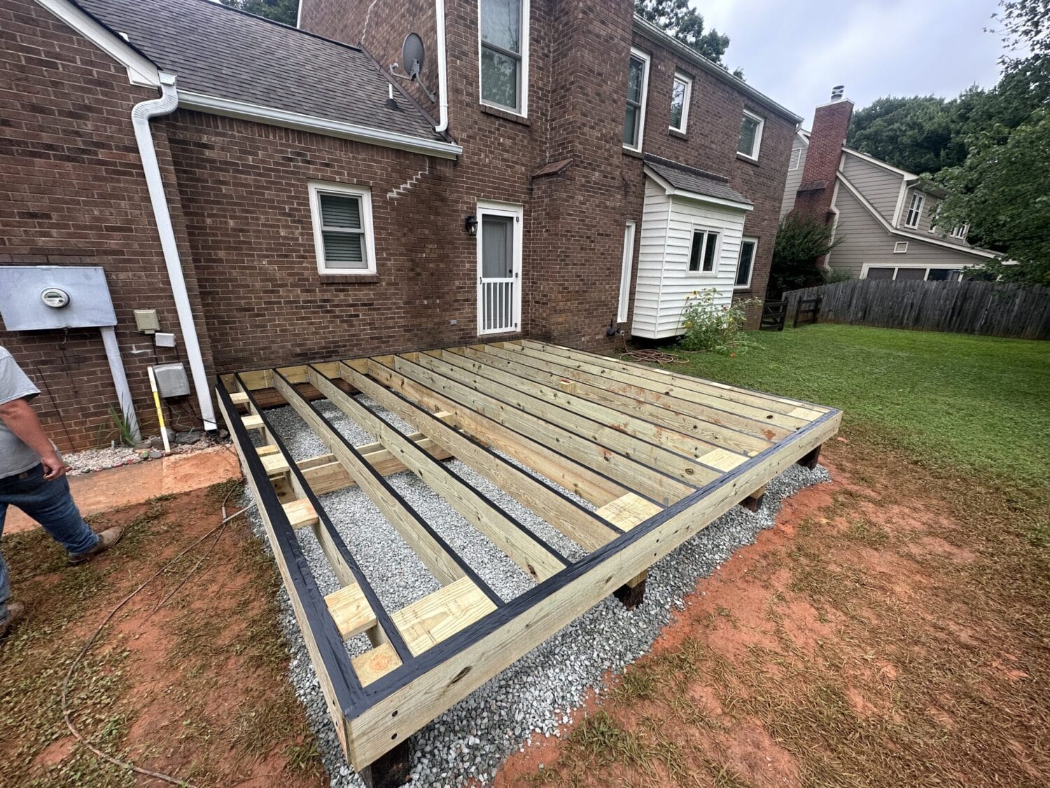 Wooden deck framing under construction attached to a brick house, with exposed beams over gravel and a partially visible worker on the left.