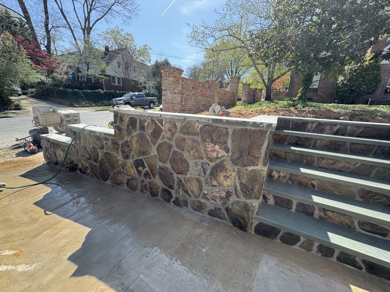 Stone retaining wall and outdoor staircase with wet pavement and construction tools visible on a sunny day in a residential neighborhood.